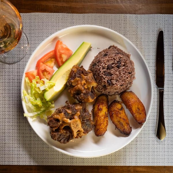 Oxtail stew with rice and beans, fried plantains, avocado, and salad on a white plate.