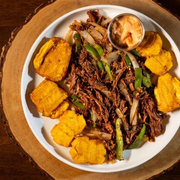 Shredded beef with green peppers, onions, fried plantains, and a side of sauce on a white plate.