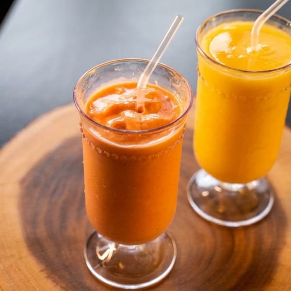 Two vibrant orange and yellow smoothies with straws, served on a rustic wooden slab.