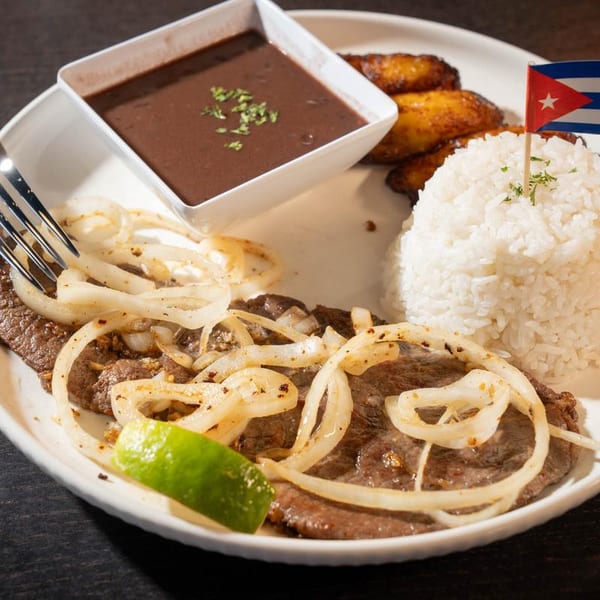 Cuban steak with onions, rice, black beans, plantains, and a Cuban flag toothpick.