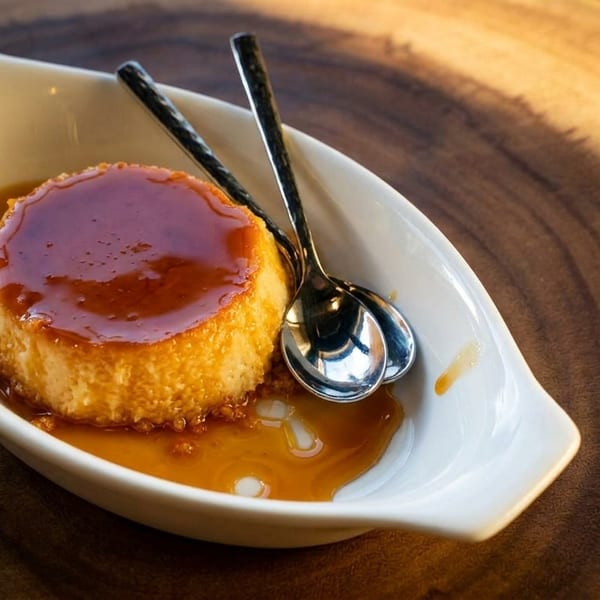 Flan dessert with caramel sauce in a white dish, with two small spoons, on a wooden surface.