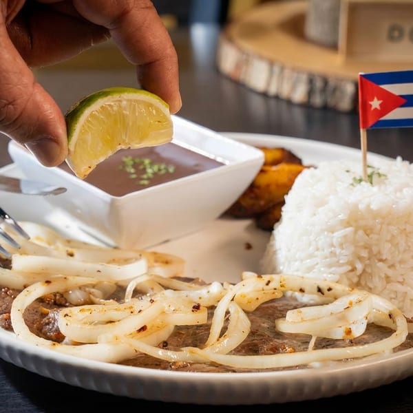 Hand squeezing lime over Cuban steak with onions, rice, plantains, and a Cuban flag.